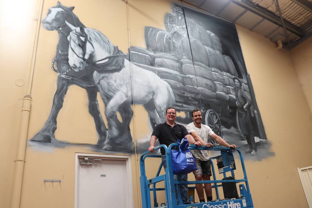 Spearwood Wool director and co-founder Andrew Basire (left) and Fremantle-based artist Jacob Shakey Butler in front of the partly completed 25 square metre mural one of two Mr Butler is painting on the walls of the show floor at Spearwood Wools Forrestdale woolstore. This mural depicts a wool wagon leaving the old Fremantle Woolstore heading to the wharf. A second mural will depict a sheep being shorn with blade shears.